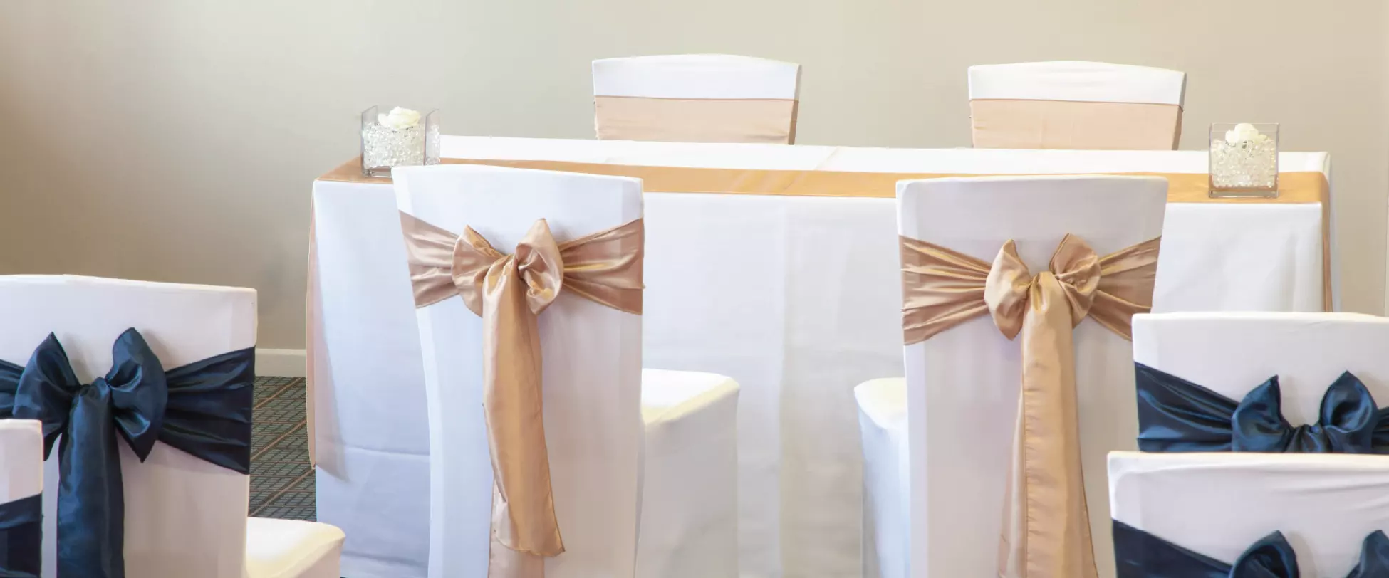A table set up for a wedding ceremony with white table and chair covers, and gold and blue ribbons.